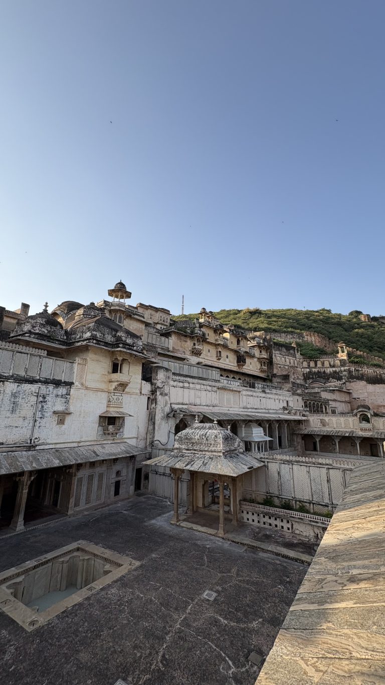 Historic white stone palace complex with ornate domes, arches, balconies, and courtyards, set against a green hillside beneath a clear blue sky.
