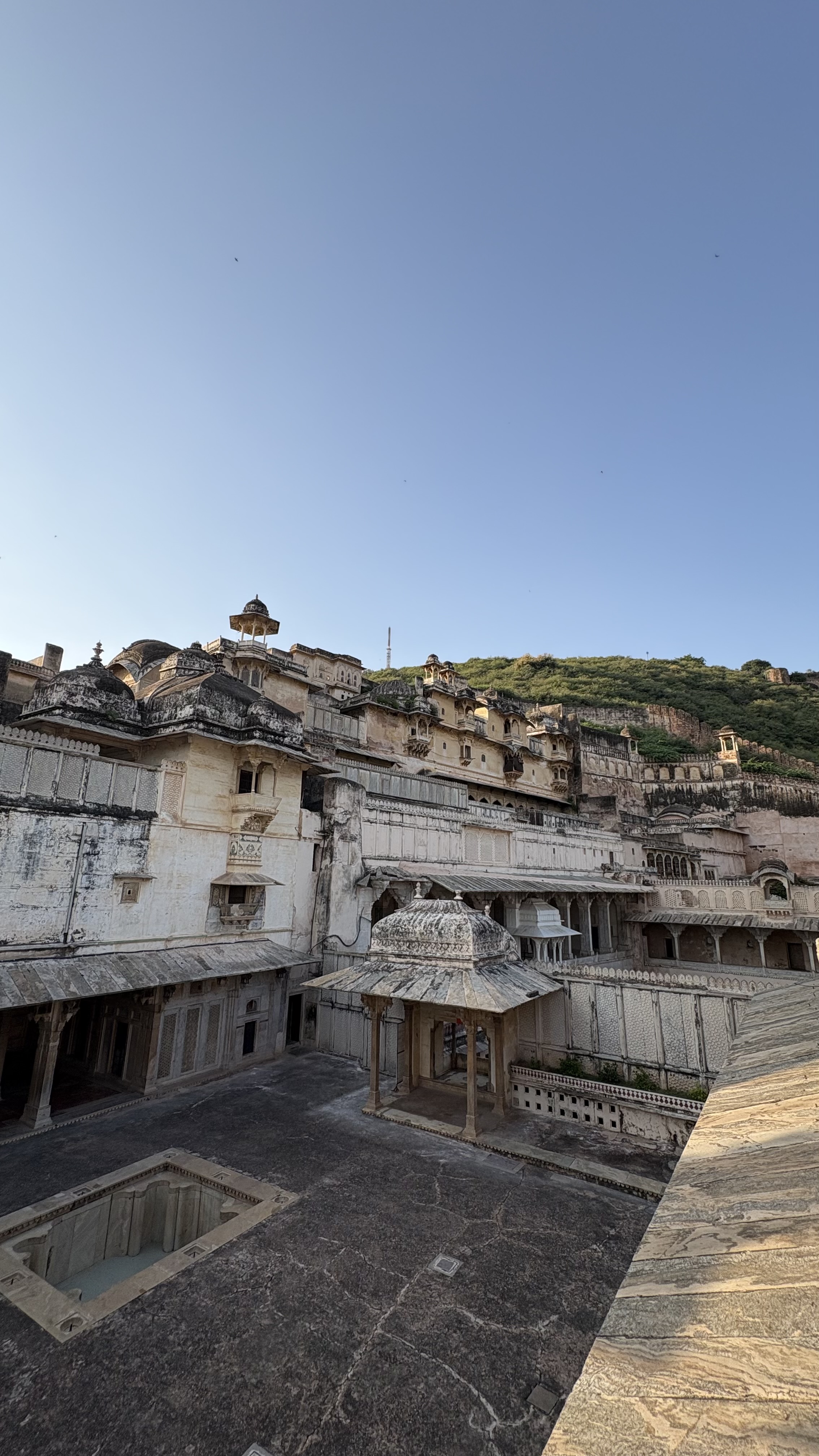 Historic white stone palace complex with ornate domes, arches, balconies, and courtyards, set against a green hillside beneath a clear blue sky.