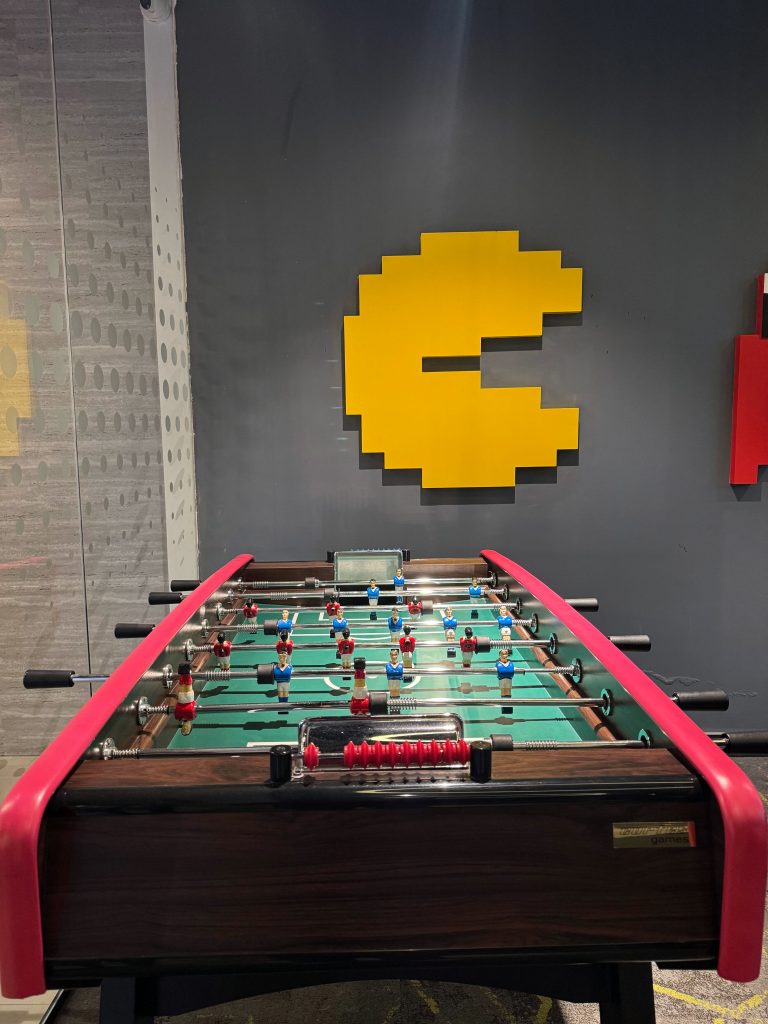 Eye-level indoor shot of a foosball table with a green playing field and black and wooden trim in the foreground, featuring a grey wall in the background adorned with a large yellow pixelated Pac-Man wall art piece.