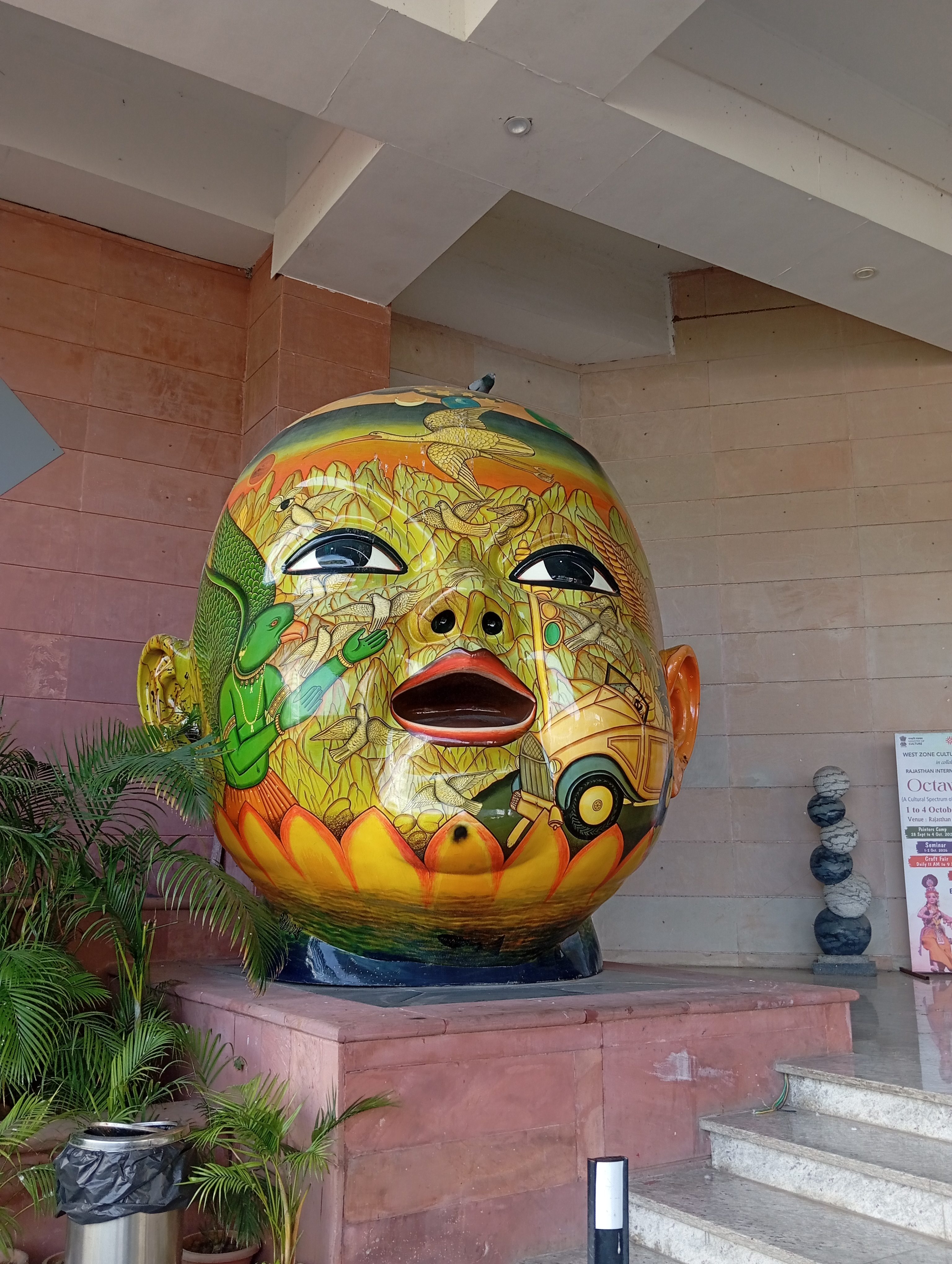 Large colorful sculptural artwork of a human head covered in intricate illustrations of birds, animals, plants, and cultural motifs, displayed on a pedestal in an indoor entrance area.