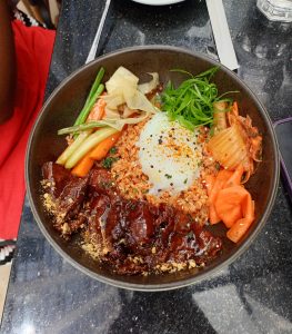 A ceramic bowl of seasoned fried rice topped with sweet soy glazed beef slices, a poached egg, shredded green onions, pickled carrots, and kimchi.