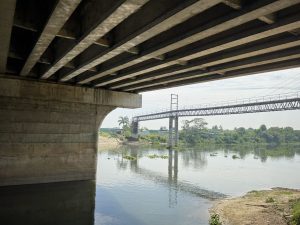 A bridge spans a river, with a larger bridge visible in the background against a clear sky.