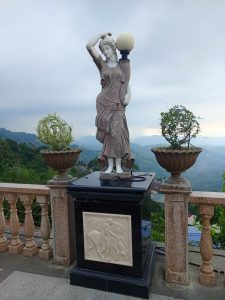 A stone garden statue of a woman draped in a flowing gown holding a round globe lamp on her shoulder, positioned on a pedestal overlooking mountain ridges.