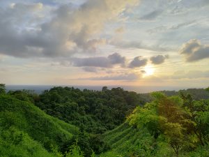 The setting sun glows through dramatic clouds above lush green forested hills, with the sea visible as a faint line on the far horizon.