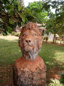 A hand-sculpted clay bust of a male lion with a detailed mane mounted on a rough wooden pedestal outdoors. 