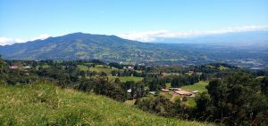 Green mountainous landscape with pastures, farms, and a distant city under a clear blue sky.
