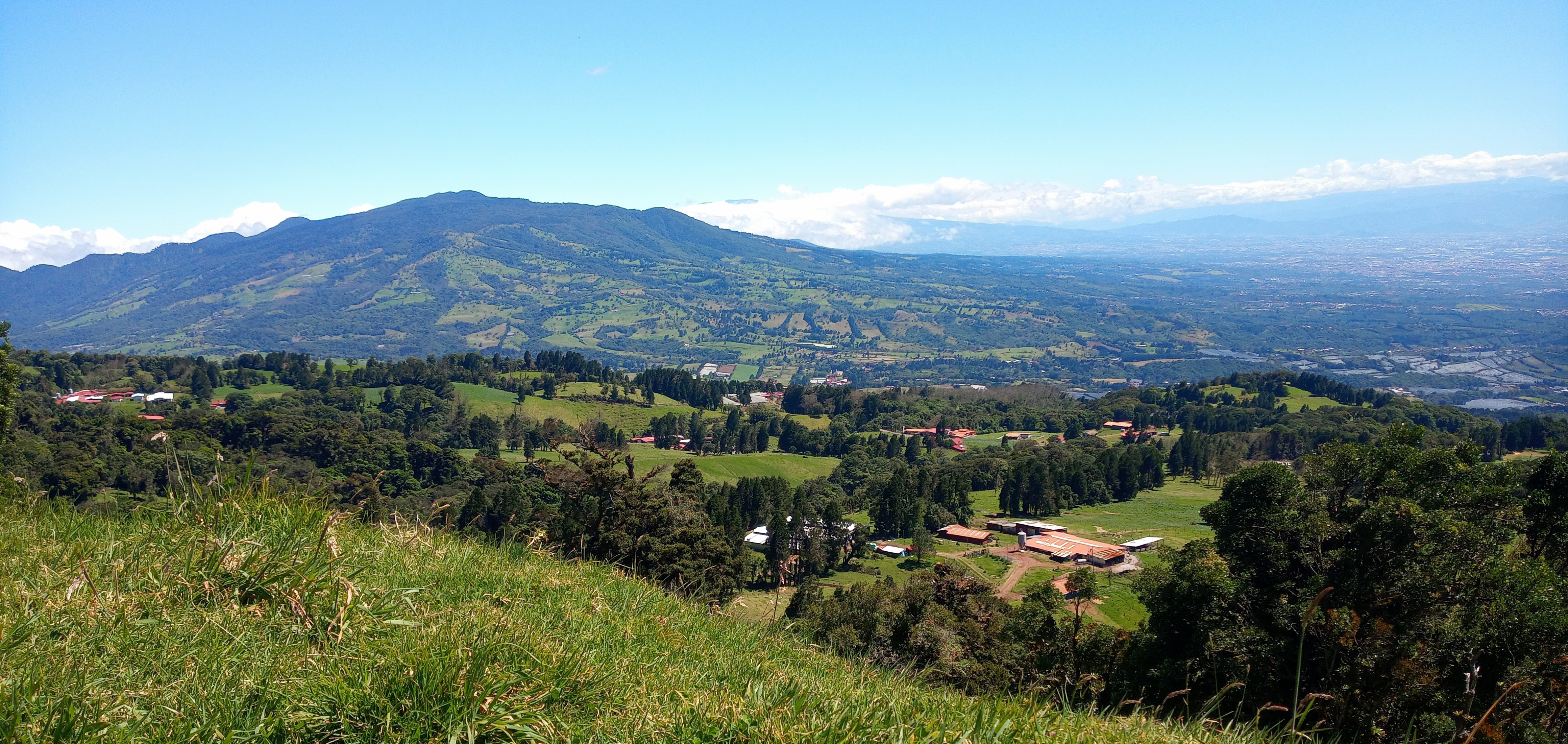 Green mountainous landscape with pastures, farms, and a distant city under a clear blue sky.