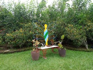 A decorative metal garden bench featuring a backrest shaped like a butterfly with colorful wire accents, surrounded by potted plants on green lawn grass.