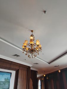 Ornate golden chandelier with crystal details hanging from a white ceiling in an elegant wood-paneled interior.