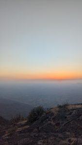 A small red sun sets over a hazy valley, casting a warm orange glow across the horizon above a rocky hillside.
