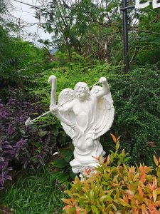 A white garden statue of a Cupid drawing a bow and arrow nestled among dense purple and green shrubbery in a garden.