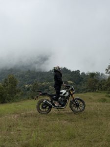 A person wearing a black outfit and colorful helmet standing beside a motorcycle in a grassy area surrounded by trees and hills under a cloudy sky.