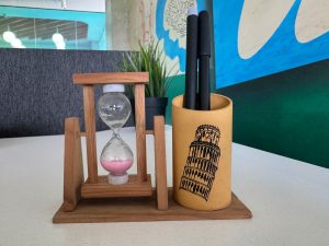 Wooden hourglass with pink sand beside a wooden pen holder on a desk.