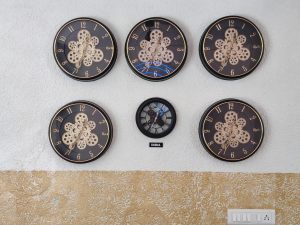 Six decorative wall clocks with dark faces and gold hands on a white and golden wall, with an “INDIA” label.