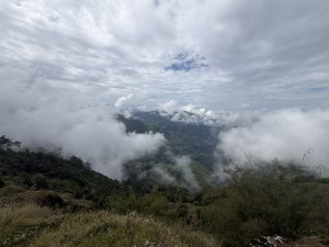 A high-altitude, panoramic view looking down over rolling, cloud-veiled mountain valleys with scattered rural settlements nestled among green terraced hillsides.