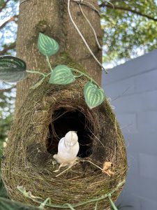A woven bird’s nest with a small white decorative bird attached to a tree trunk.
