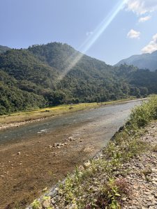 The Adhikhola River, bathed in the golden glow of the scorching sun,its waters shimmering under the clear blue sky with magestic hills and crop fields in the background .