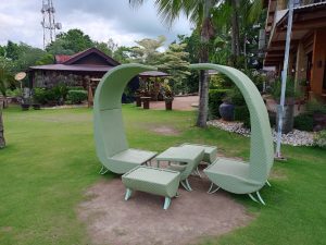 A light green woven wicker outdoor lounge furniture set with arched canopy armchairs and low matching tables placed on a manicured lawn.
