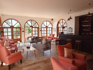 A bright, spacious indoor lounge with peach armchairs, grey tufted sofas, and a dark wood bar counter facing large arched doors with water views.