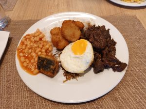 A hearty Filipino cuisine breakfast platter featuring savory cured beef tapa, fried chicken nuggets, baked beans in sauce, a sunny-side-up egg, and white rice.