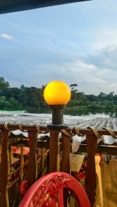 A round yellow glowing lamp mounted on a wooden balcony railing overlooking a calm river and distant trees.