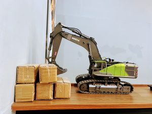 A toy Huina excavator model on a wooden surface with its bucket resting next to small cardboard boxes. 