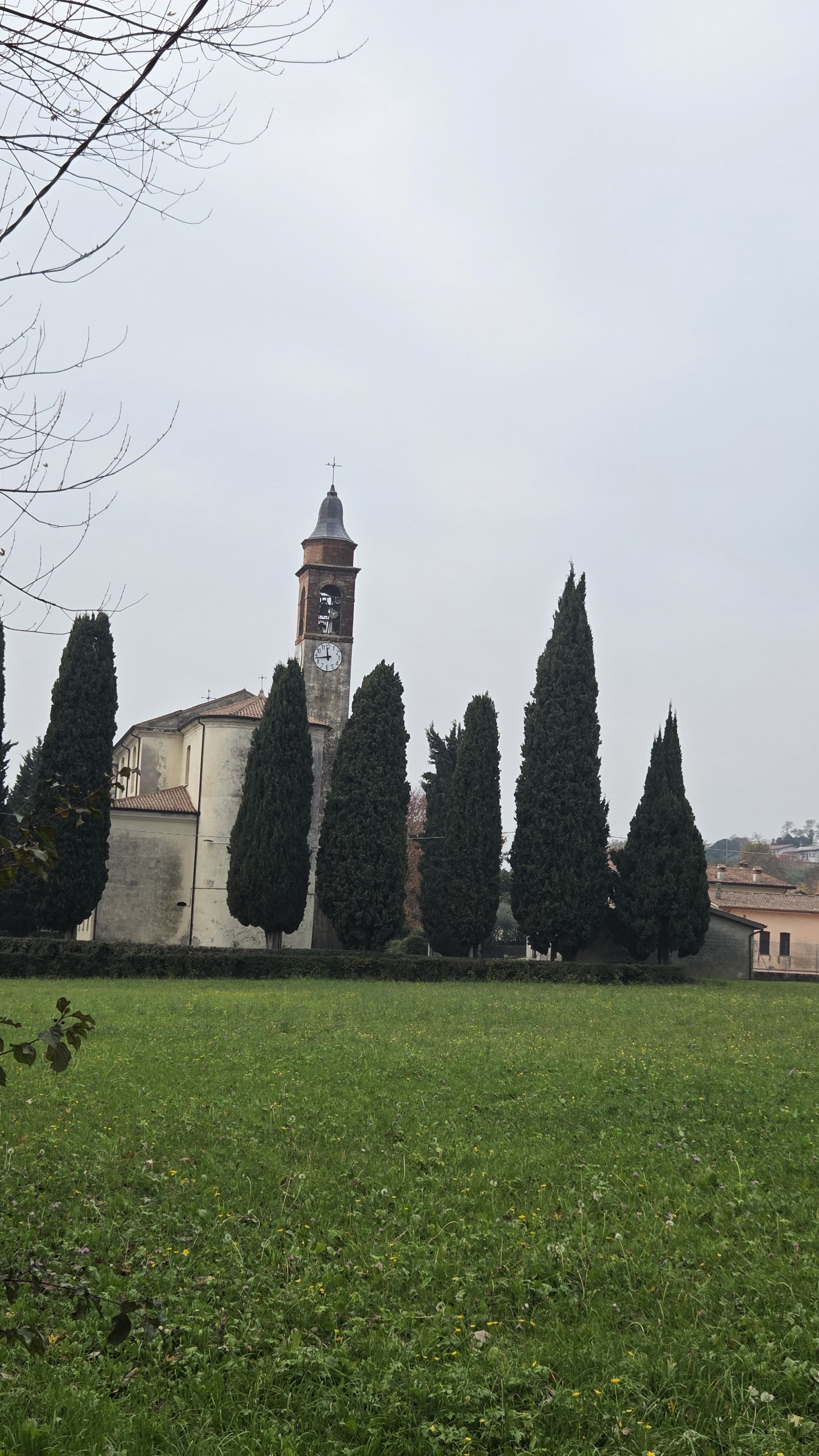 A tall stone bell tower with a prominent clock face and a dark conical roof topped with a cross. A neat row of tall, slender, dark green cypress trees stands in front of the cream-colored stone church building.