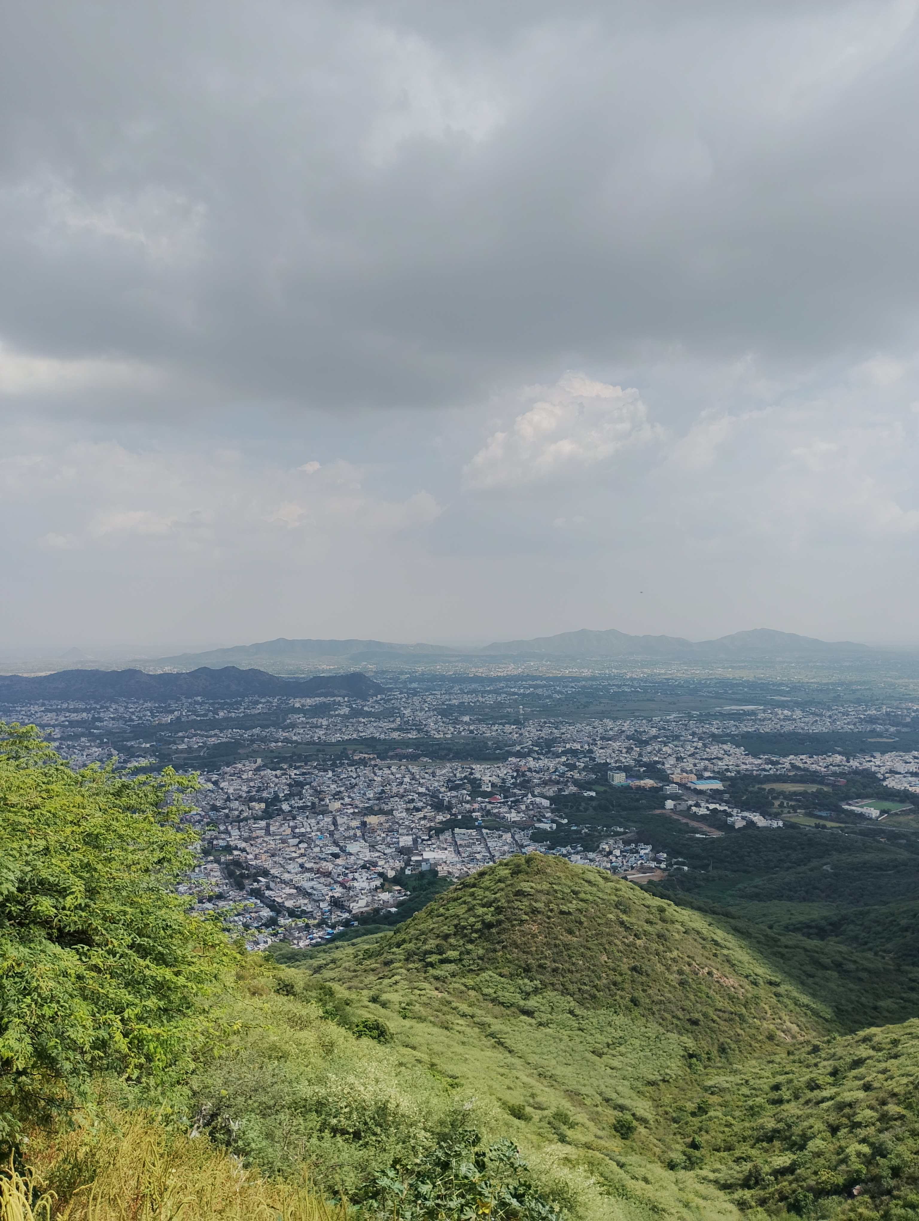 Panoramic view of a sprawling city surrounded by lush green hills and distant mountains beneath a cloudy sky.