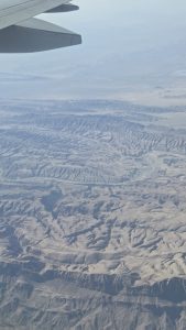 An aerial view of a rugged desert with deep canyons and mountain ridges stretching to a hazy horizon.