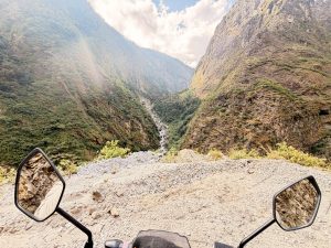 A wide point-of-view perspective from behind motorcycle handlebars, looking out over a steep gravel ledge down a massive, tree-lined mountain valley with a rushing stream flowing through the canyon bottom under a partly cloudy sky.