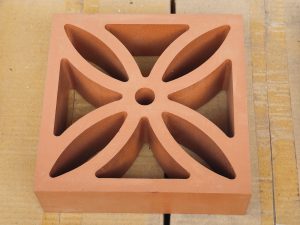 A terracotta architectural ventilation block featuring a symmetrical floral pattern with eight curved openings. The warm clay color and geometric design are clearly visible. Location: Perumanna, Kozhikode, Kerala.