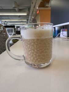 Close-up shot of a textured glass mug filled with foamy, creamy coffee or milk tea, sitting on a white office desk with an office interior and computer monitor softly blurred in the background.
