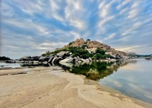 Granite rock formations rise above a calm river, their warm, earthy colors reflected in the water. A dramatic, cloud-filled sky and the broad riverbank create a scenic landscape showcasing the natural beauty of the Hampi region of Karnataka.