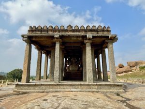 A stone pavilion with multiple pillars shelters a seated Ganesha statue at Hampi's historic ruins. The structure stands on a rocky landscape beneath a blue sky with scattered clouds. Location: Sasivekalu Ganesha Temple, Hampi, Karnataka.