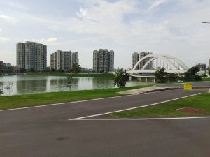 A white arched bridge spanning Nirjhor Lake in Dhaka Cantonment, Bangladesh, with high-rise residential buildings, green lakeside banks, and a paved road beneath a partly cloudy sky.