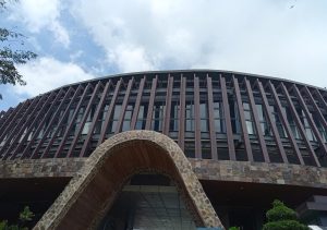 A modern curved open rooftop building at the top of Cebu highlands featuring vertical brown slats and an organic stone archway entrance beneath a blue sky.
