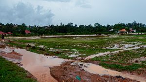 Wide-angle view of a grassy field and dirt path heavily flooded with muddy brown rainwater under an overcast grey sky, with houses and lush green trees visible in the background.