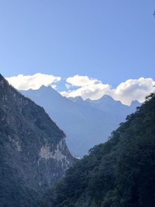 scenic mountain gorge framed by dark, forested slopes, revealing sharp, sunlit mountain ridges and puffy white clouds in the distance.