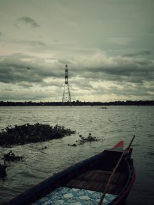 A small wooden boat on a rippling river with floating plants, a metal tower, and cloudy skies.