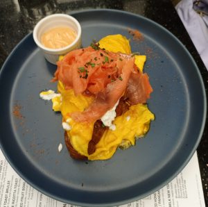 A breakfast dish featuring soft scrambled eggs and delicate folds of smoked salmon atop a toasted croissant, served with a side of creamy spiced sauce.