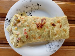 A savory chicken supreme burrito filled with vegetables and corn, coated in a creamy herb sauce and served on a patterned ceramic plate.