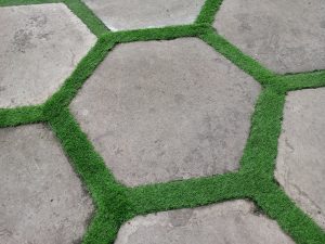 A stone patio pavement pattern featuring large hexagonal concrete slabs bordered by bright green strips of artificial grass.