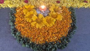 Flower rangoli made of yellow and orange petals, with a lit diya in the center.
