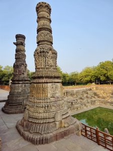 all, intricately carved stone pillars rise beside the stepped tank of the Sun Temple. The detailed sculptures, blue sky, and green water highlight the beauty of this historic architectural site. Location: Sun Temple, Modhera, Mehsana district, Gujarat.
