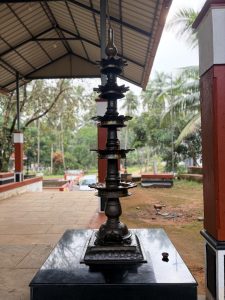 A tall, multi-tiered traditional brass oil lamp (Sthambavilakku) stands on a black stone pedestal in an open-air temple pavilion in Kerala.