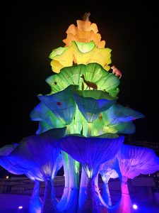 A towering nocturnal light installation at night shaped like a layered floral tree, illuminated in glowing rainbow gradients of blue, green, yellow, and orange.