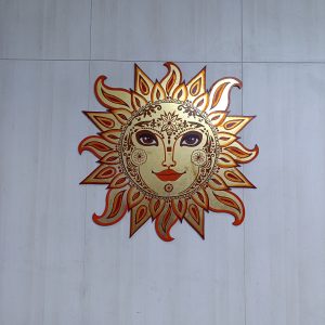Decorative Indian-style sun artwork featuring a detailed human face surrounded by ornate golden and red sun rays, mounted on a tiled wall.