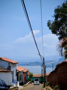 A lake with mountains behind, there is a street with houses and a car in the middle on the street