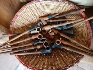 A woven circular wicker basket containing a collection of traditional smoking pipes with ceramic clay bowls and long wooden stems.