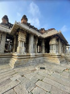 An intricately carved stone pavilion showcases ornate pillars, sculpted figures, and detailed granite reliefs. Warm sunlight highlights the craftsmanship of this historic Vijayanagara-era architecture against the blue sky. Location: Hampi, Karnataka.
